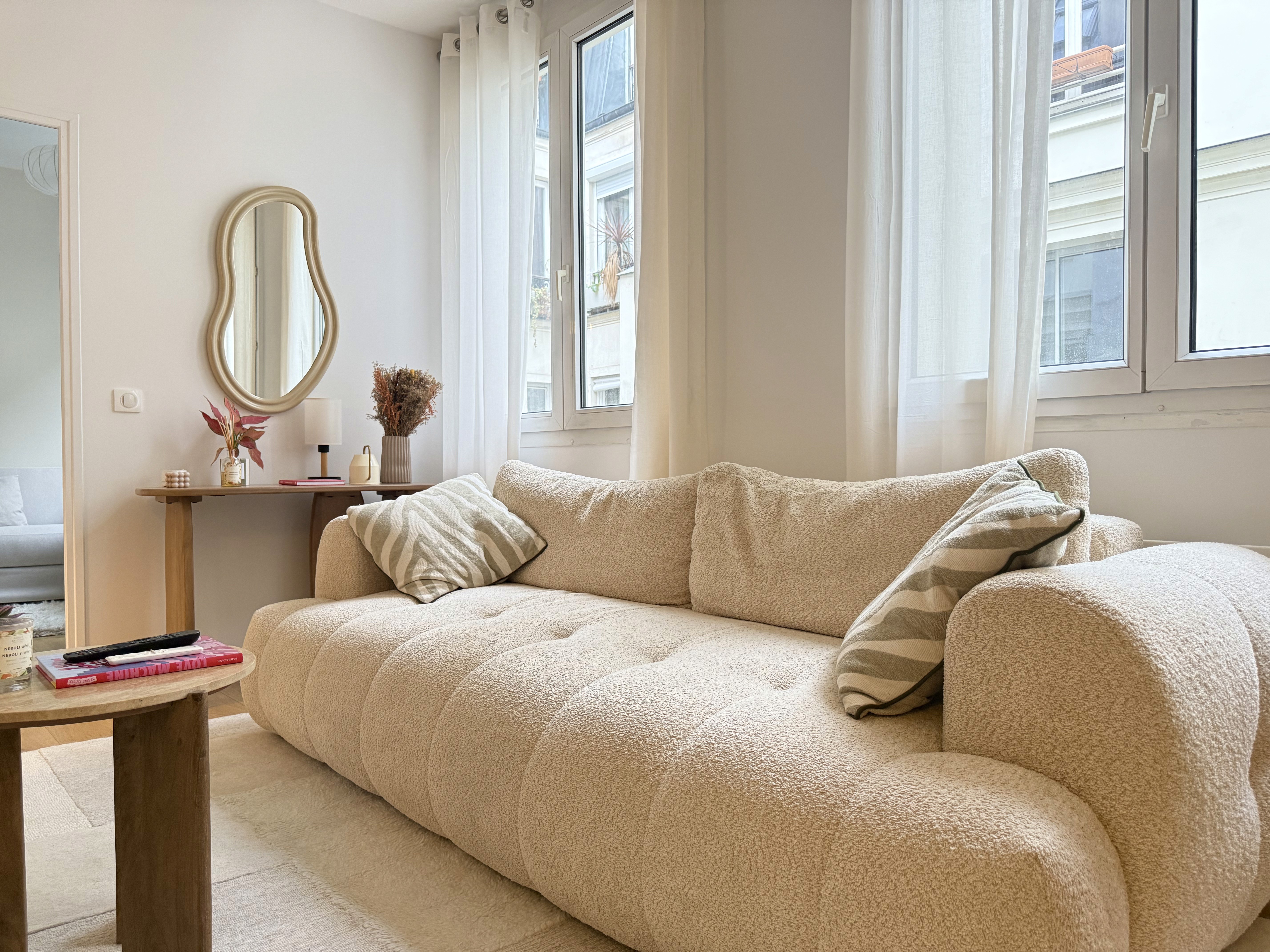 Salon parisien lumineux avec un grand canapé beige en tissu bouclé, parquet en bois clair et miroir décoratif organique.