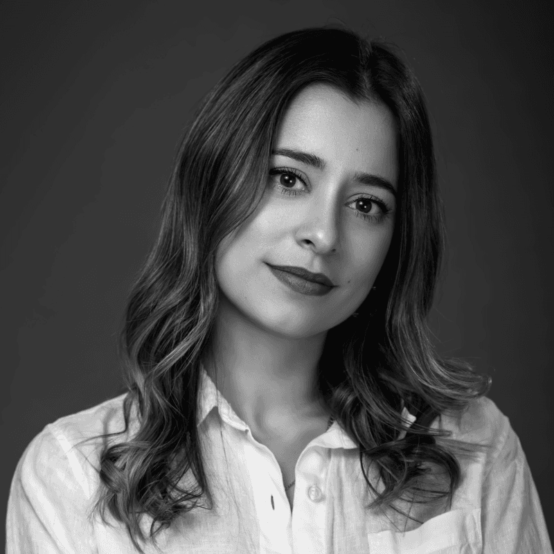 A woman with long, dark hair and a slight smile poses in front of a bright yellow background. She is wearing a black top and a necklace.