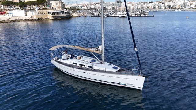 Aerial starboard profile view of the Dufour 40 Performance sailing yacht at anchor off the Malta waterfront, showing the clean white hull with dark blue topsides, teak deck, tan bimini, blue carbon running backstay, and the Sliema harbour development and marina packed with yachts in the background