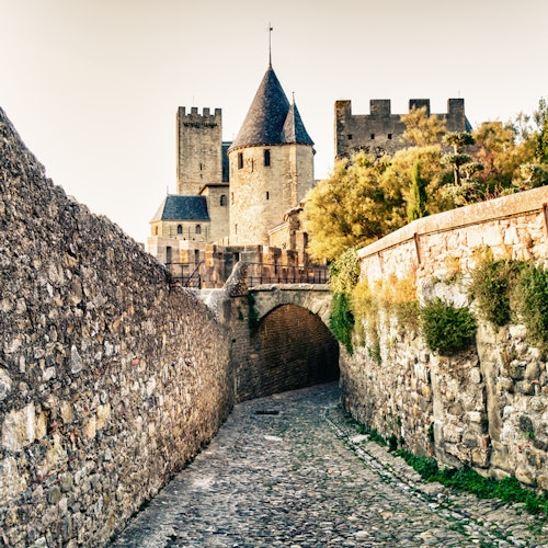 A cobblestone path leads to a historic stone castle with multiple towers and a bridge, flanked by high stone walls.