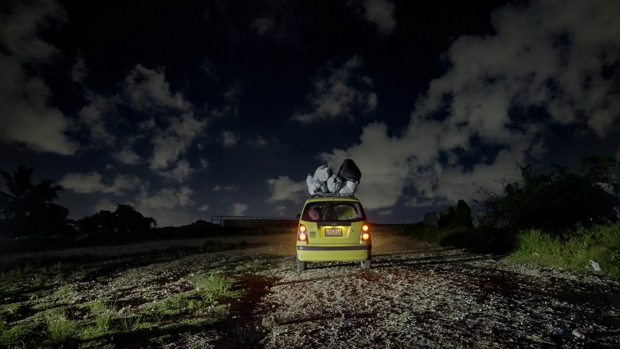 A small yellow car is driving on a gravel road at night, its trunk open and overflowing with large bags. The sky is dark, filled with scattered clouds, and the headlights of the car illuminate the road ahead.