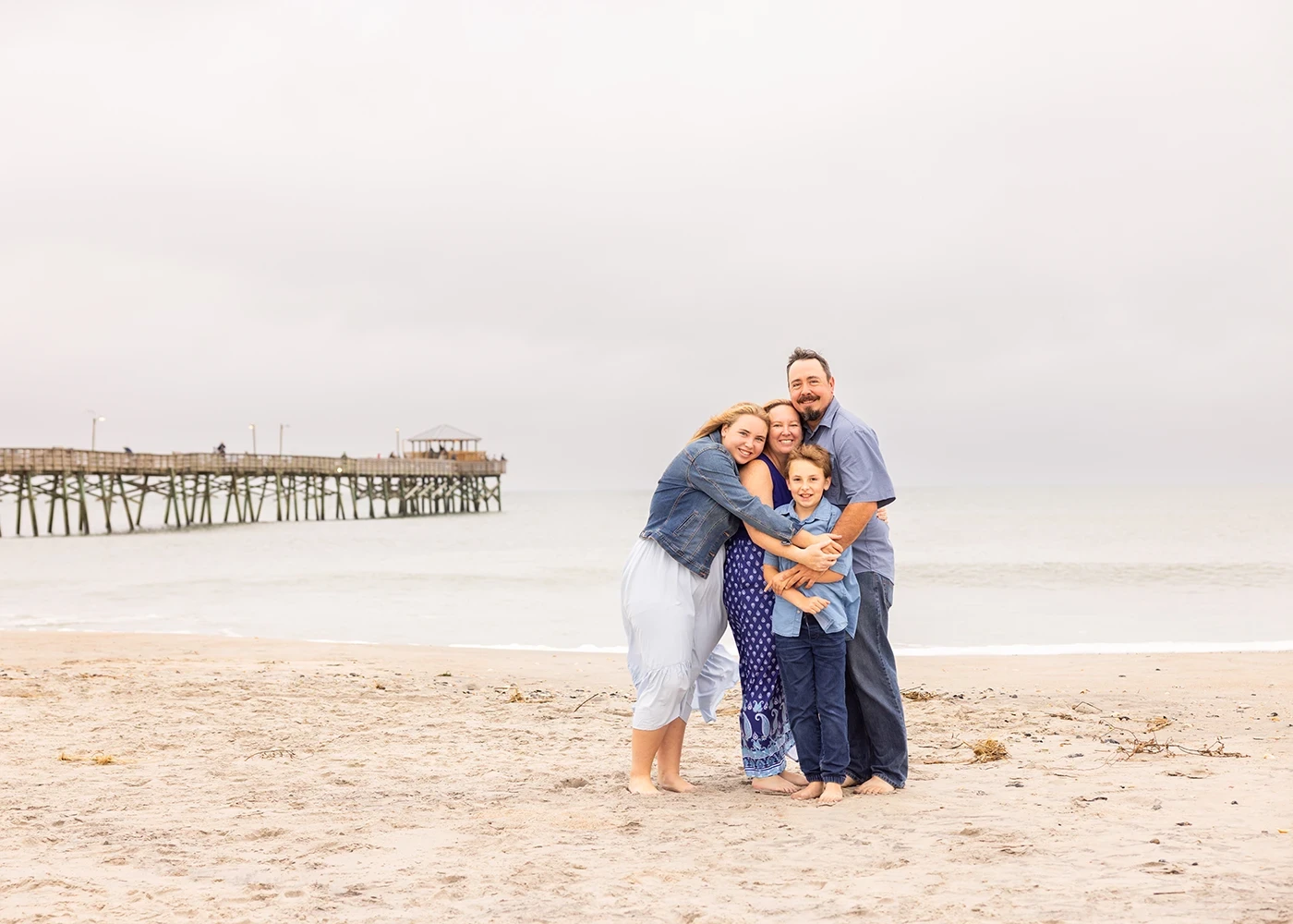 Family of four hugging on the beach near a pier