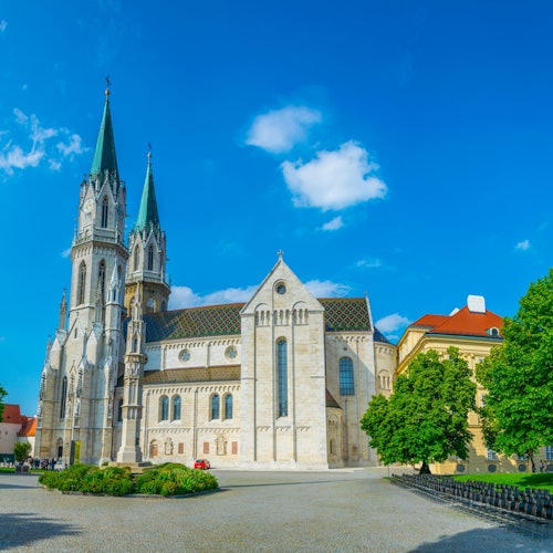 A stone cathedral with two tall spires next to a yellow building and green trees under a bright blue sky with white clouds.