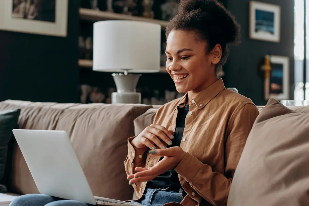 A woman sits on a sofa using a laptop, smiling and gesturing with her hands, with a lamp and framed photos in the background.