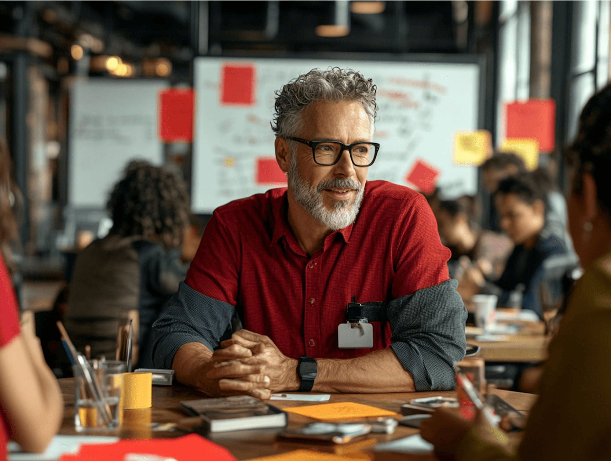 A bearded man wearing glasses and a red shirt sits at a wooden table in a busy, modern office environment with colleagues engaged in discussion and red sticky notes on a whiteboard in the background.