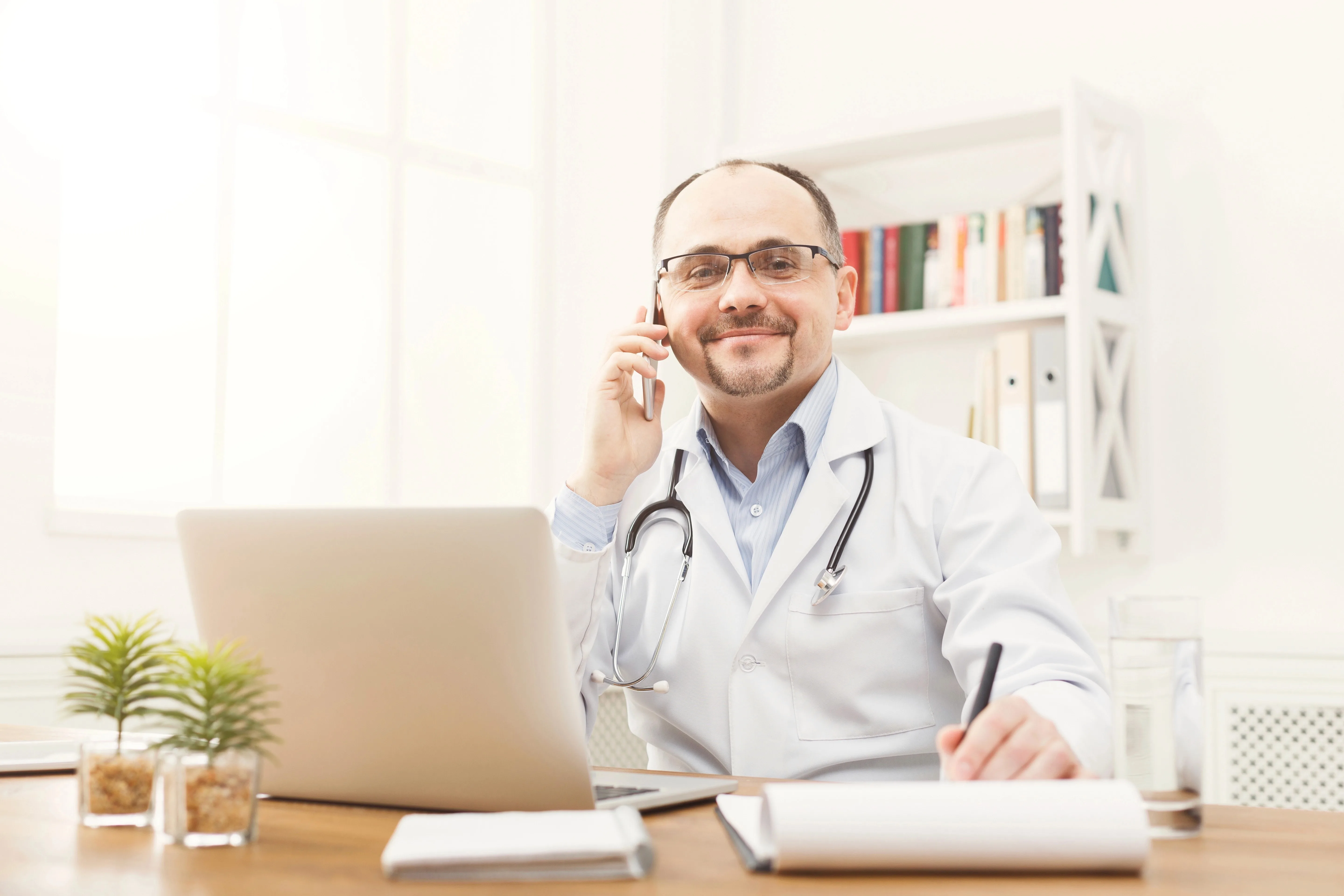 Male physician wearing a white coat with stethoscope around the neck, sitting on a chair with a table and laptop in front, one hand answering a cellphone and another hand holding a pen.