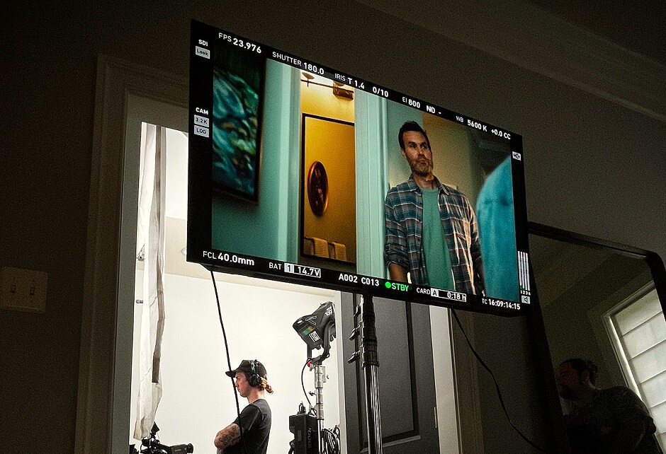 A behind-the-scenes view of a film set, showing a cameraman at work and a monitor displaying a scene with a man standing in a doorway.