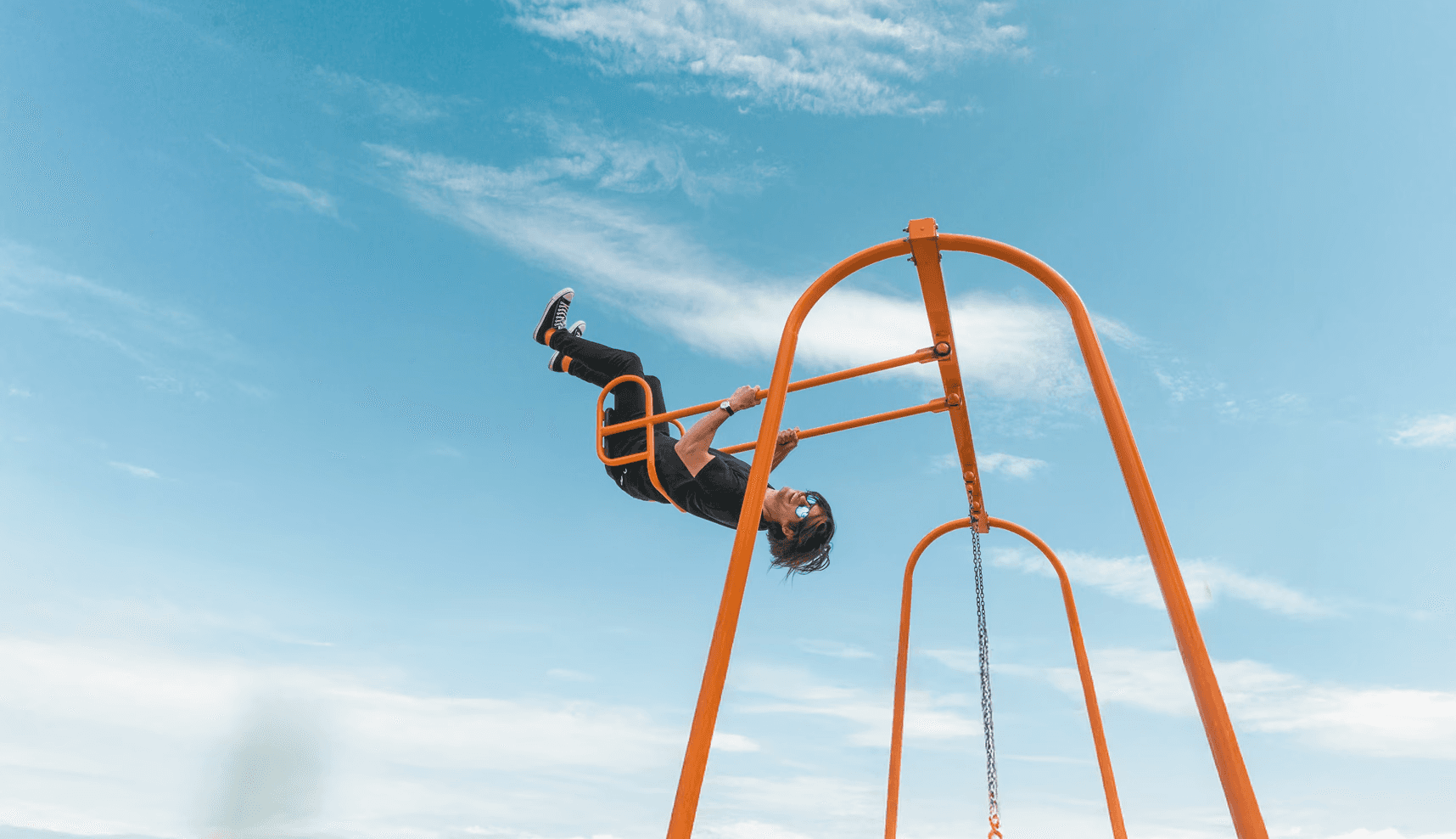 Person hanging upside down from orange playground bars against a bright blue sky.
