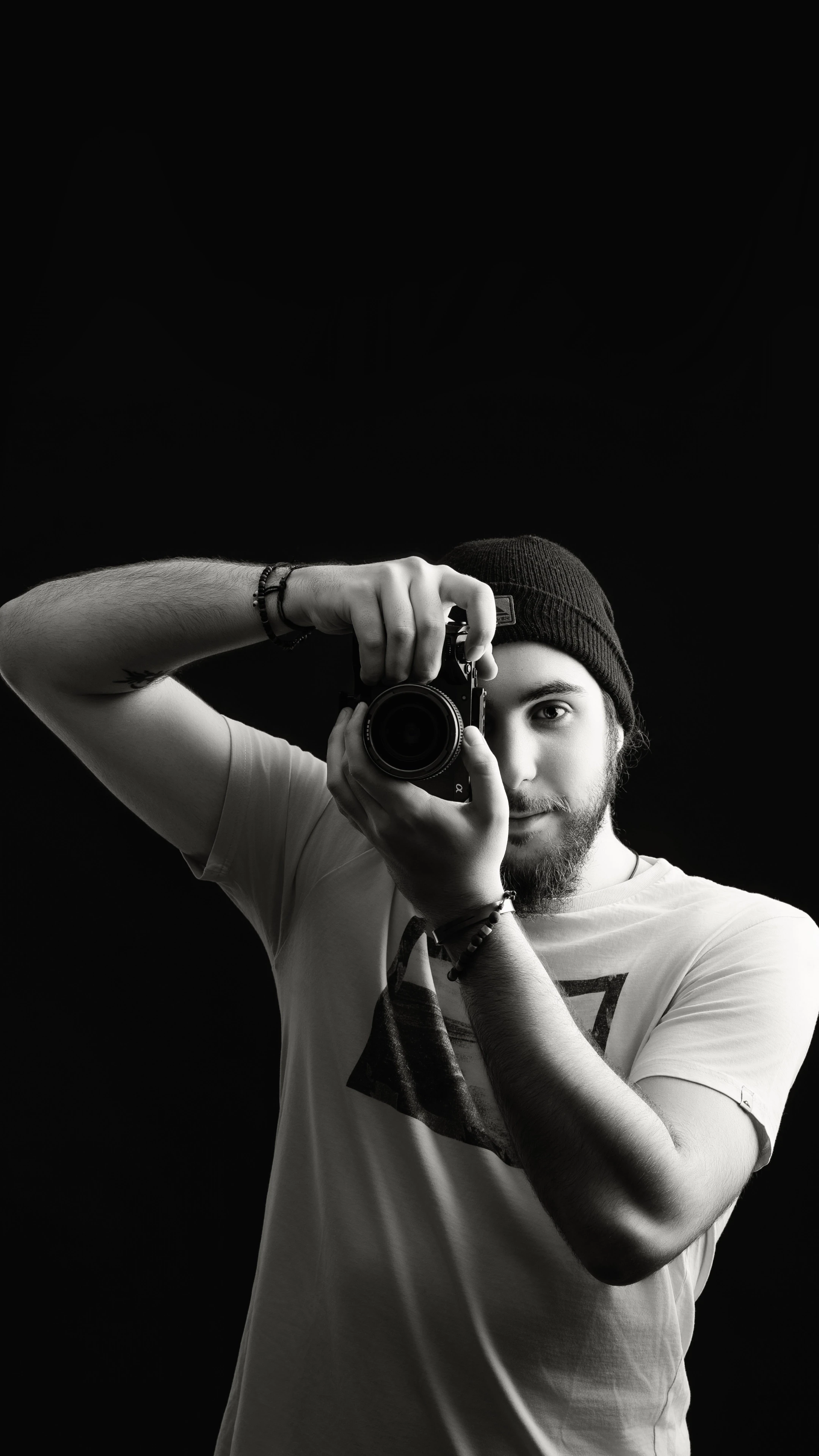 Black and white portrait of videographer holding a camera to eye, capturing a shot against a dark background, representing Raybrand Media’s professional videography, creative storytelling and visual content production