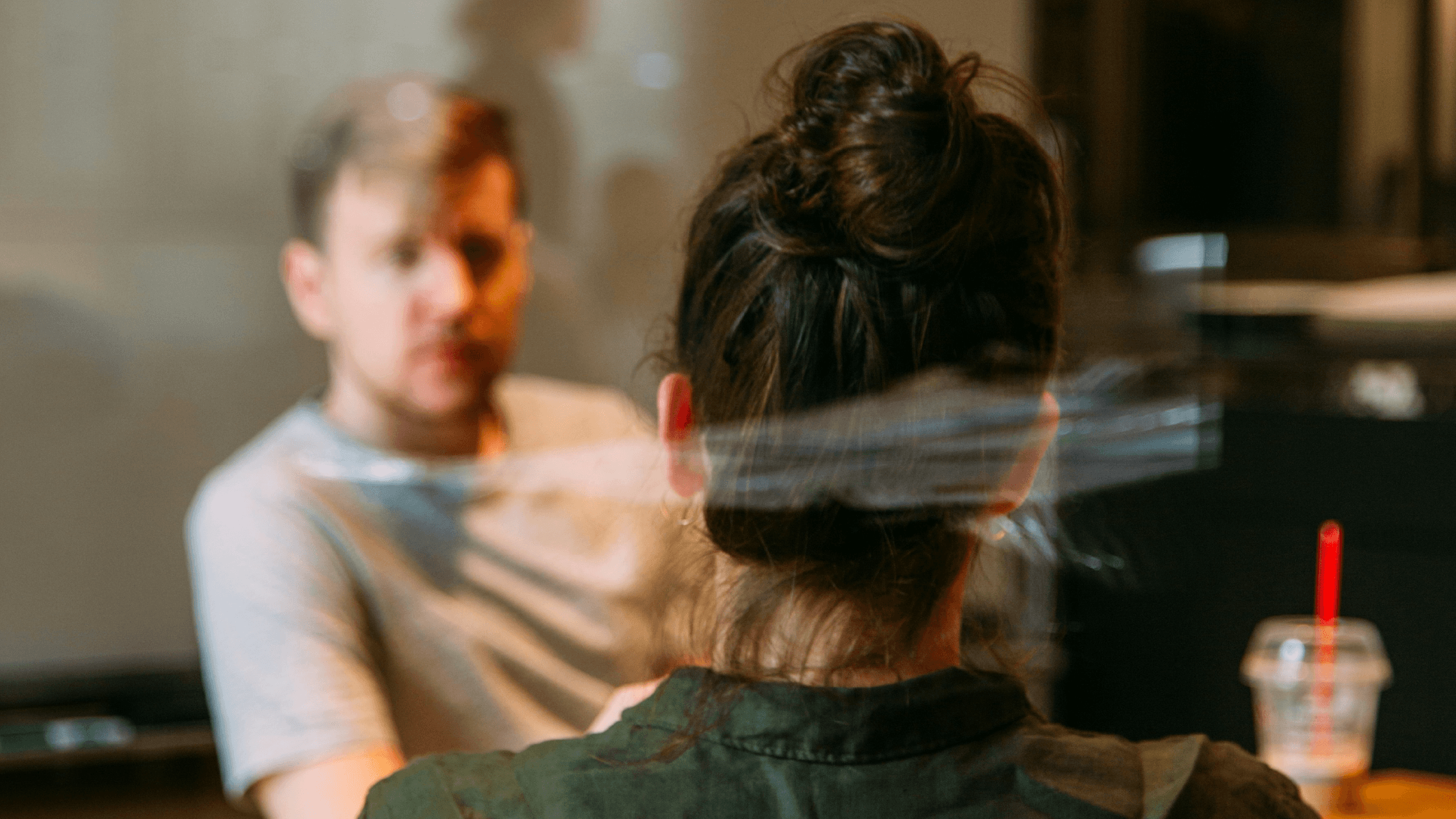 A woman viewed from behind sits across a table from an out-of-focus man during an interview.
