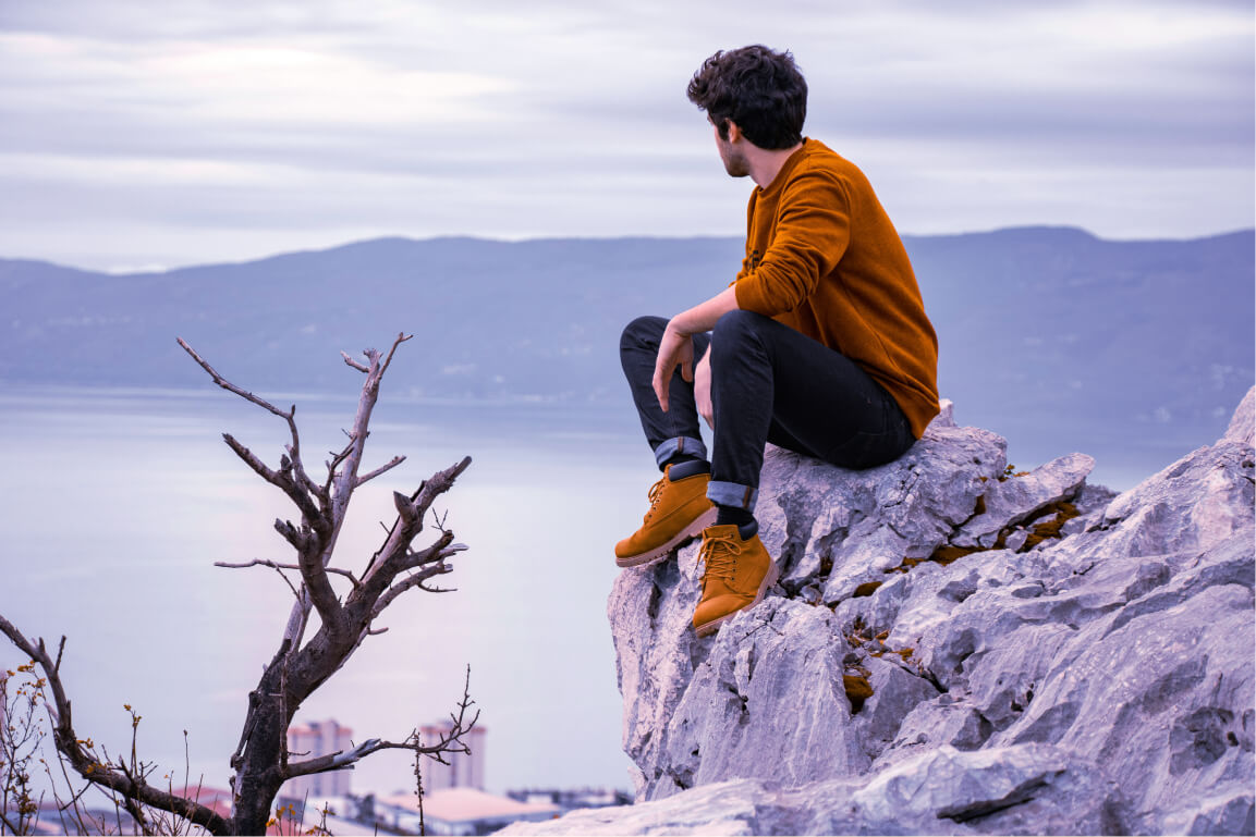 Mental health man with orange shirt blue jeans sitting on rock overlooking mountain lake