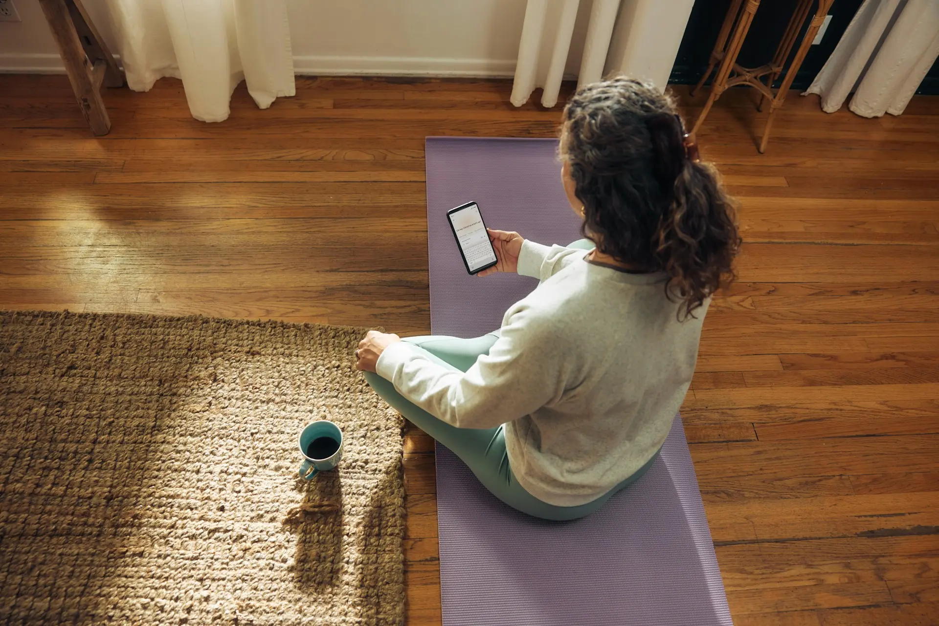 Woman sitting on a yoga mat at home using a smartphone to check class schedule or booking information.