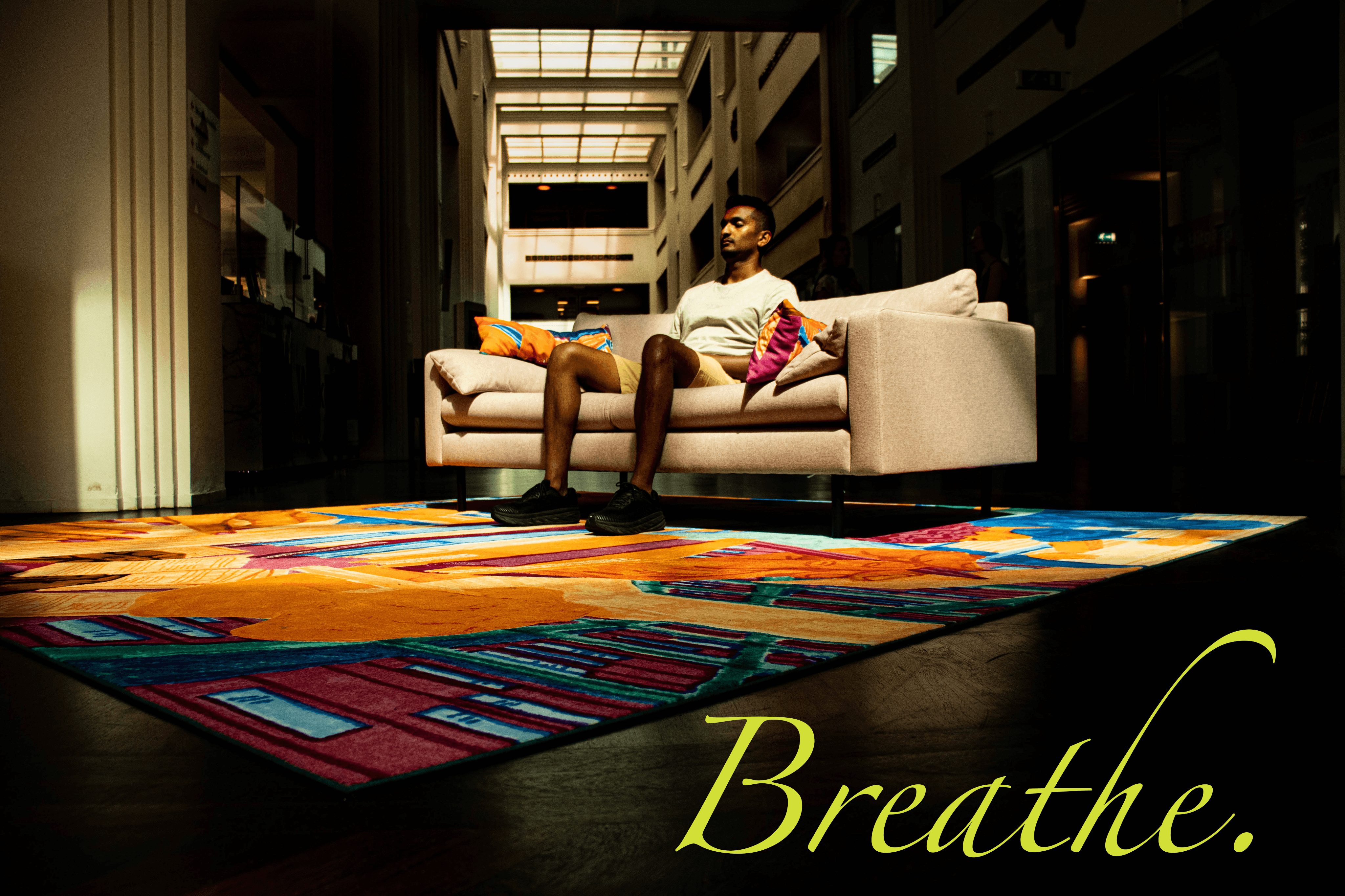 A man sitting quietly on a modern couch in a sunlit lobby, eyes closed in a mindful breathing practice, with warm light and colorful rug creating a calm atmosphere — promoting mindfulness and pain-relief meditation in Oak Park.
