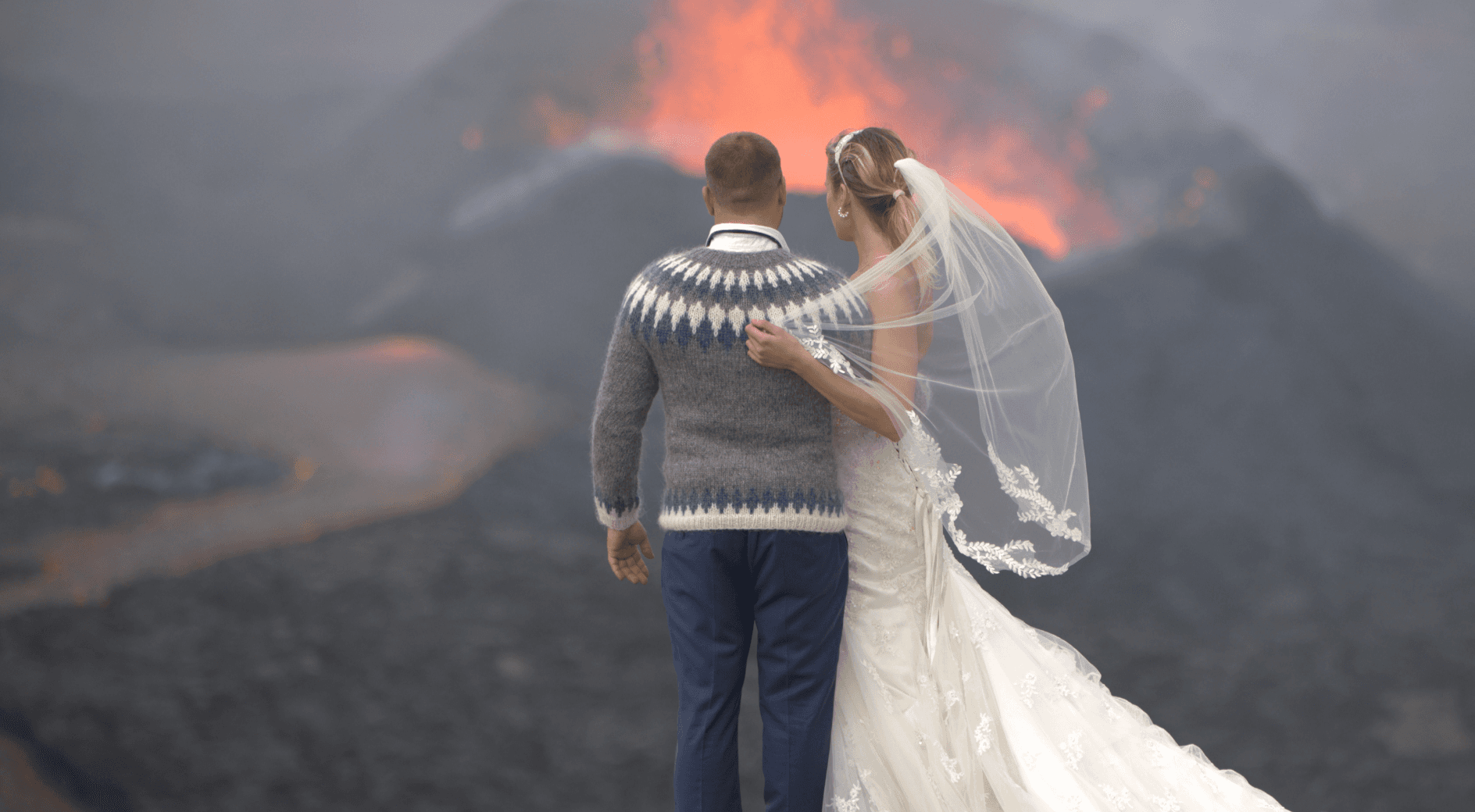 A bride and groom stand arm-in-arm facing an erupting volcano.
