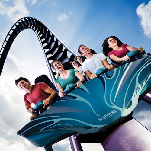 Група хора се возят с вълнение на влакче в увеселителния парк SeaWorld с примка и фоново небе, покрито с облаци.