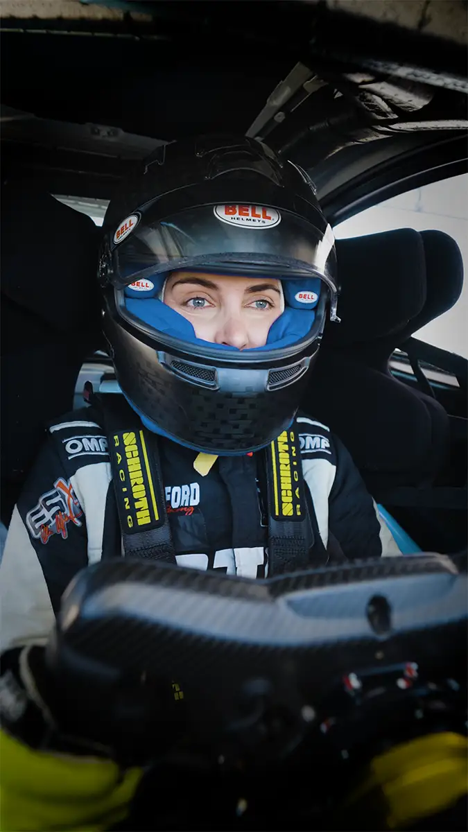 Emelia Hartford wearing a helmet and safety gear inside a race car, focusing intently ahead.