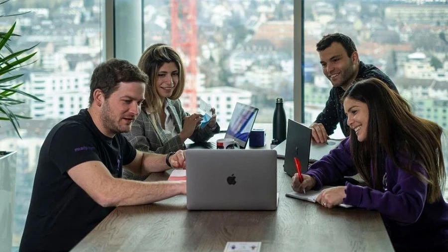 Four colleagues sitting around a table in a modern office, smiling and collaborating while looking at a laptop, with a cityscape visible through large windows in the background.
