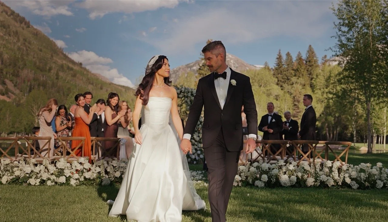 Newlyweds walk hand in hand from an outdoor ceremony lined with white flowers and mountain views.