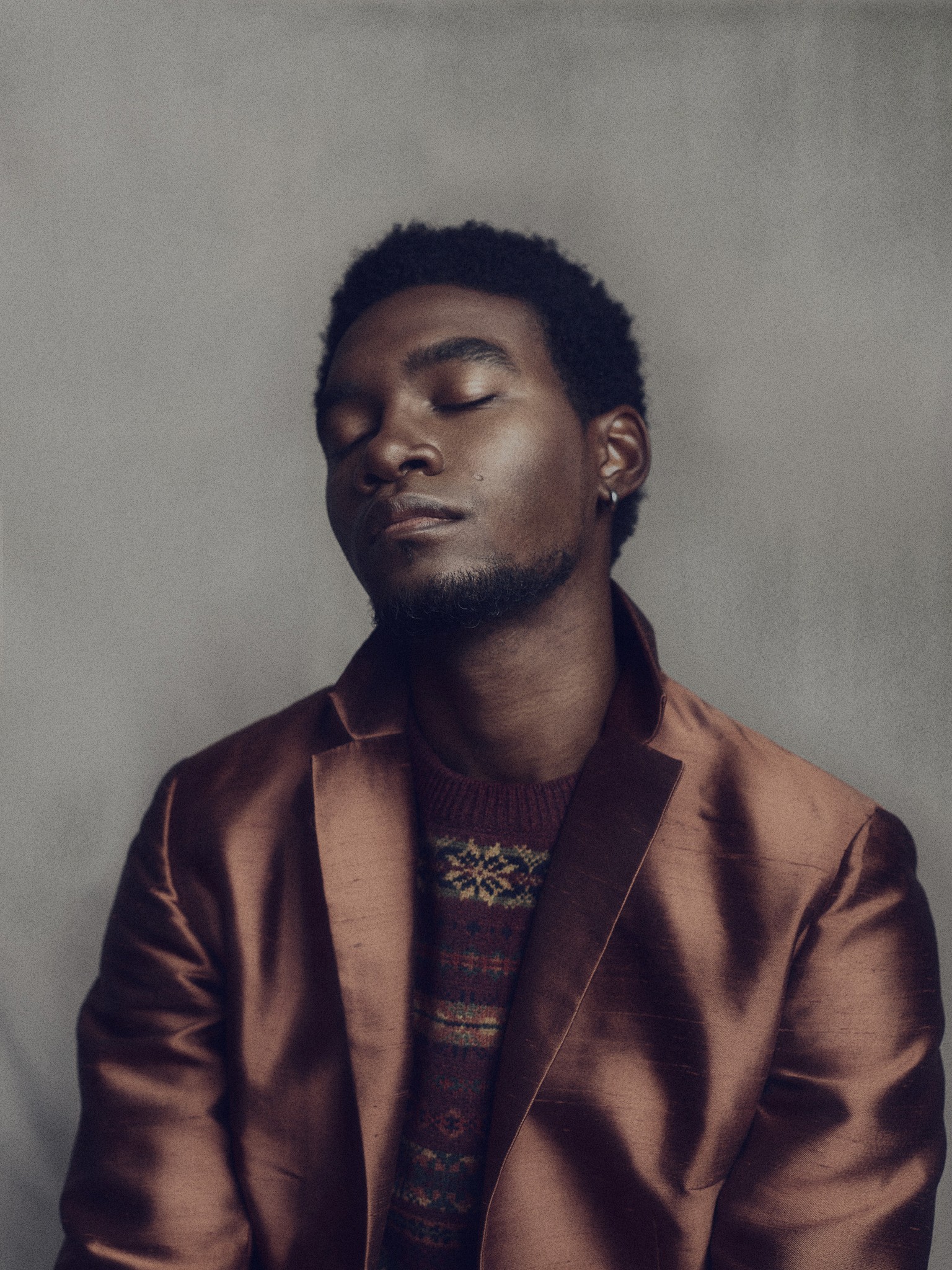 A poetic and expressive photographic portrait of a black male, wearing a silk jacket. The subject has his eyes closed in an expresive pose. 