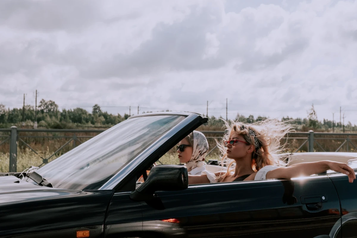 Two women riding in a convertible on an open road under a cloudy sky.