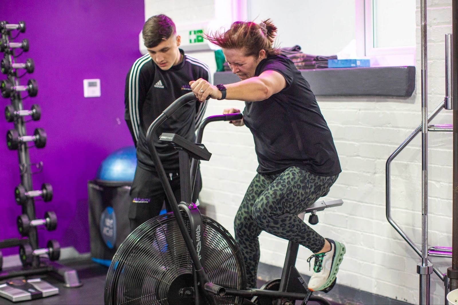 Woman using an air bike during a HIIT exercise routine session with a personal trainer.