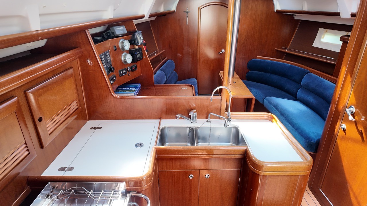 Interior saloon and galley of a Beneteau Oceanis Clipper 373, showing warm teak-finished cabinetry, a stainless steel double sink with two taps, a gimballed gas cooker with fiddle rails, and a Raymarine navigation and instrument panel. Blue velvet upholstered settee seating is visible to the right.