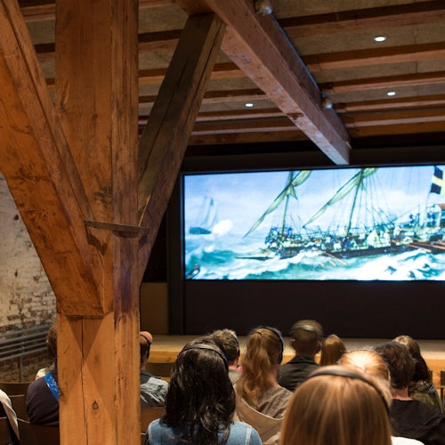 Ihmiset istuvat puuhuoneessa, katsellen suurta näyttöä, joka esittää purjelaivaa myrskyisillä merillä.
