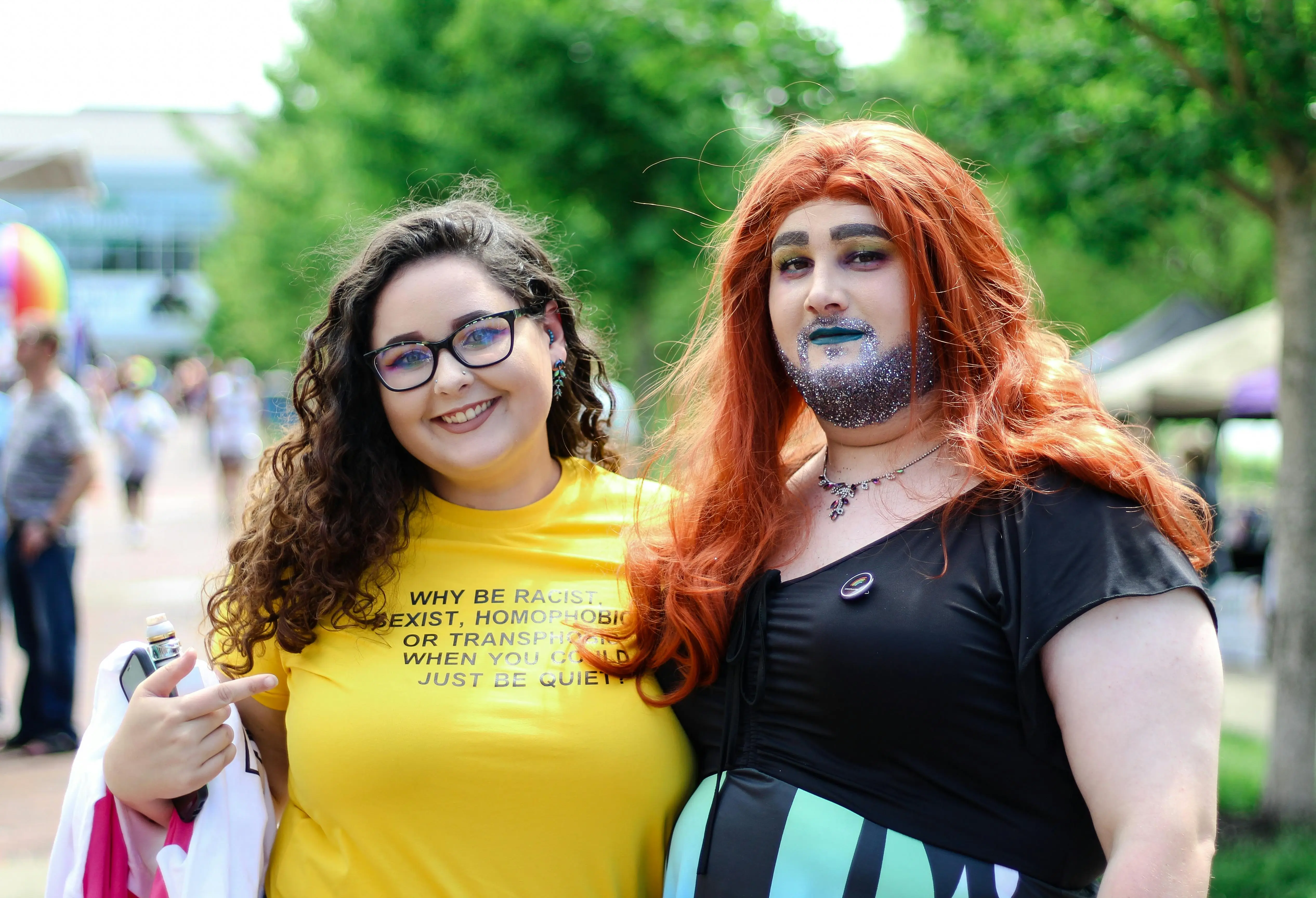 Two people are standing outside on a sunny day, one wearing a bright yellow t-shirt with an anti-discrimination message, and the other in an outfit with blue and black stripes and ginger hair, set against a background of greenery and a pathway.
