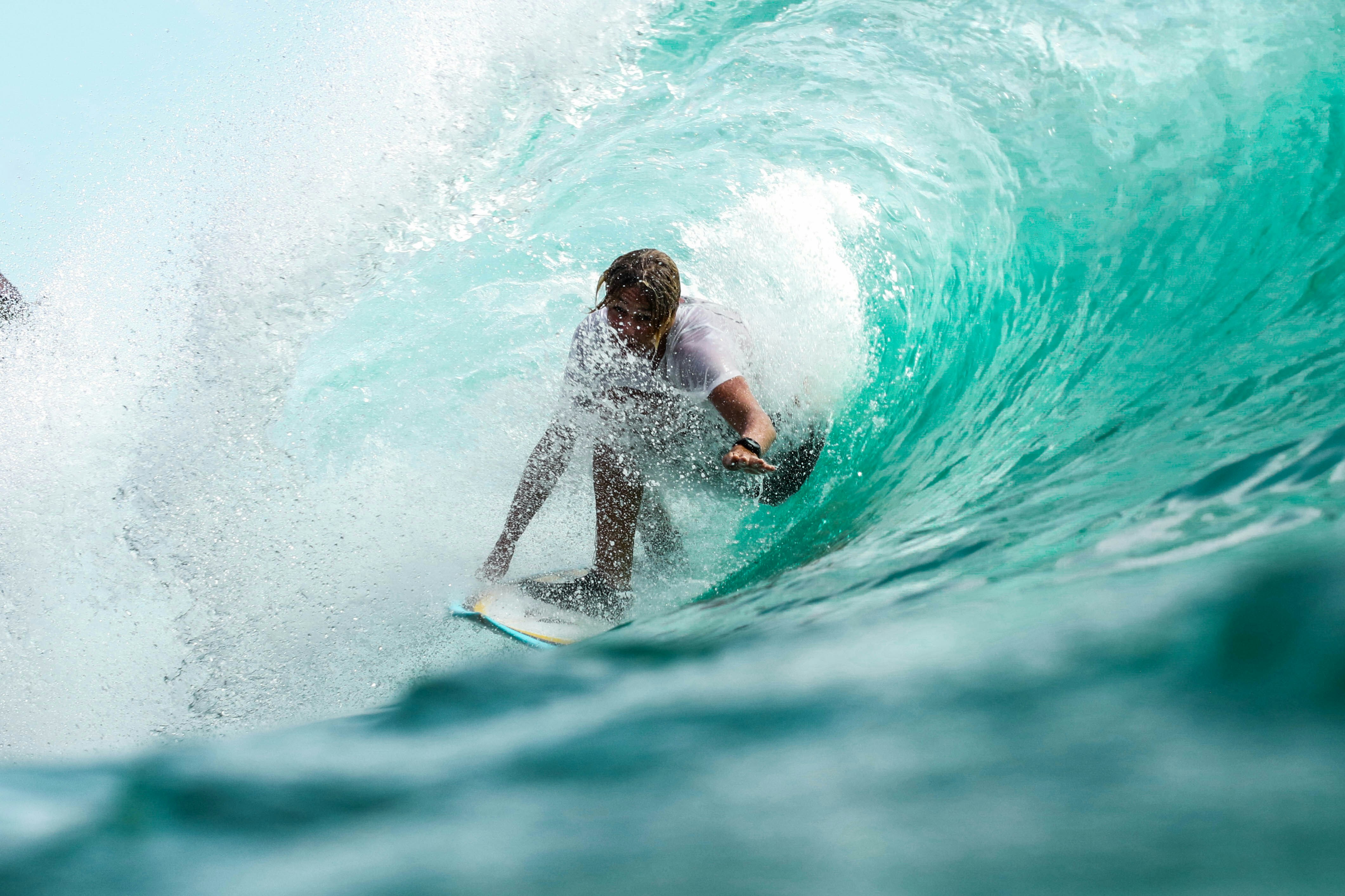 A surfer crouches low on their board while riding inside a turquoise wave, with water spraying around them as they glide through the barrel.