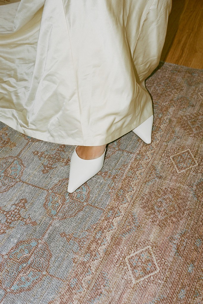 Close-up of a bride’s pointed white heels and silk wedding dress hem indoors
