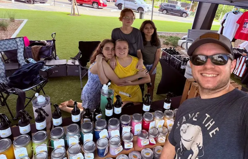 A group of people are gathered at an outdoor market stall displaying an array of jars and bottles, with a variety of goods visible on the table, under a sunny sky with grass and parked cars in the background.