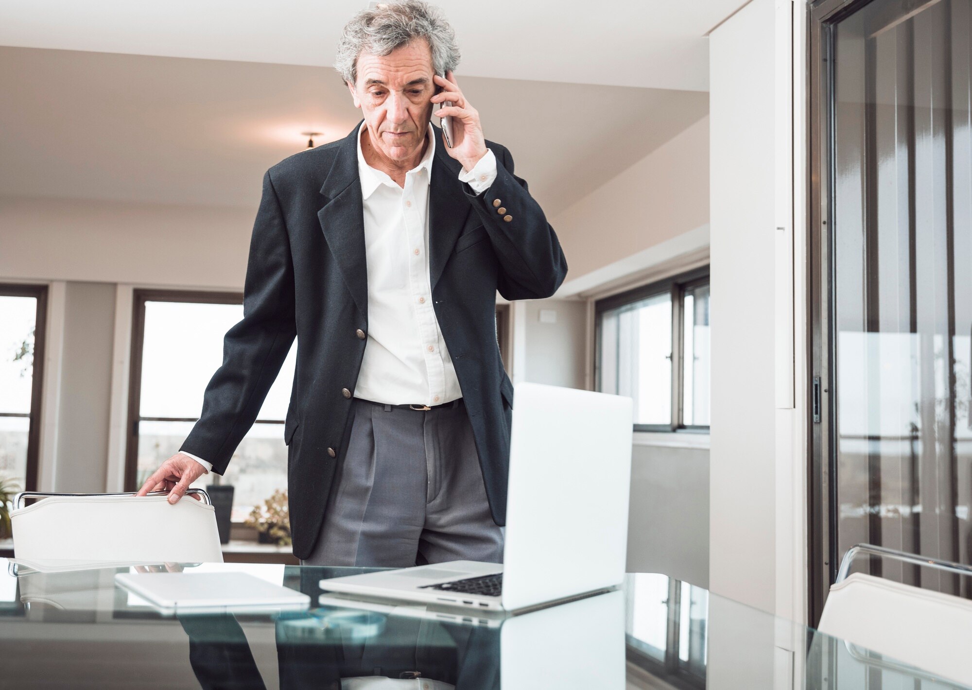 Makelaar in pak voert telefoongesprek staand achter glazen bureau in lichte moderne woning
