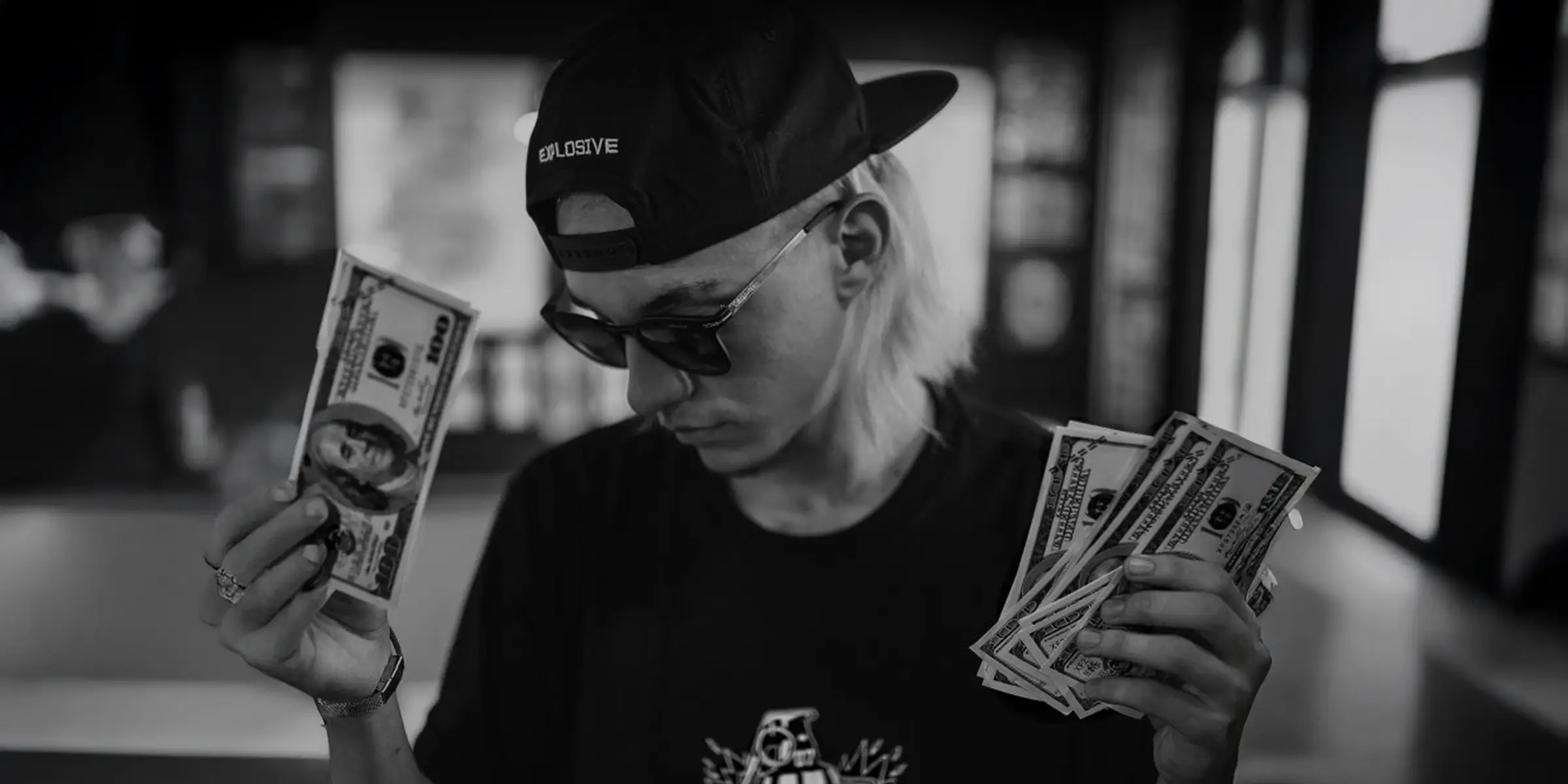 Person wearing a backwards cap and sunglasses, holding several US dollar bills in both hands and looking down at them, shot in black and white indoors.