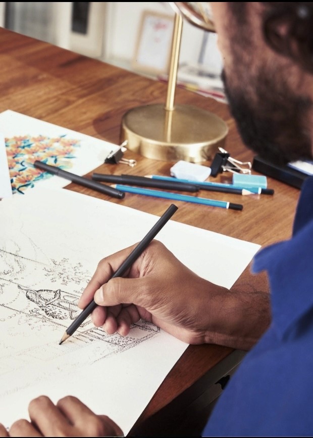 A close-up of a person's hand holding a pencil, drawing on paper with colorful art supplies scattered on a wooden table.
