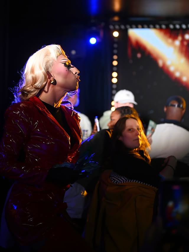 A candid shot of a drag performer with bright blonde hair and a shiny red jacket interacting with the crowd during a performance, with audience members visible under atmospheric lighting