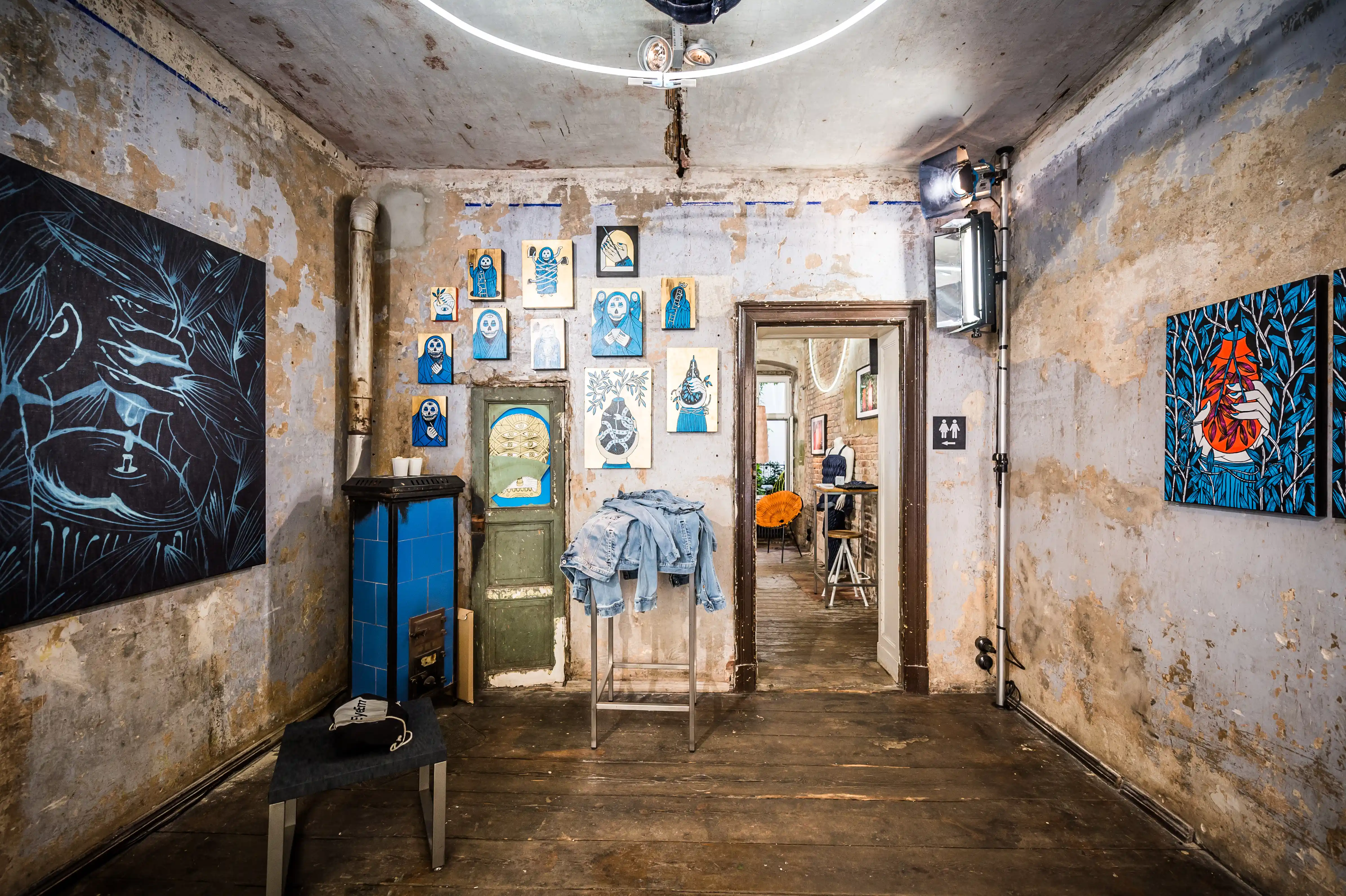 Exhibition room at the MUSTANG Street ART Lab with raw patina walls, blue artworks, a denim jacket on a stand and a vintage blue tiled stove.