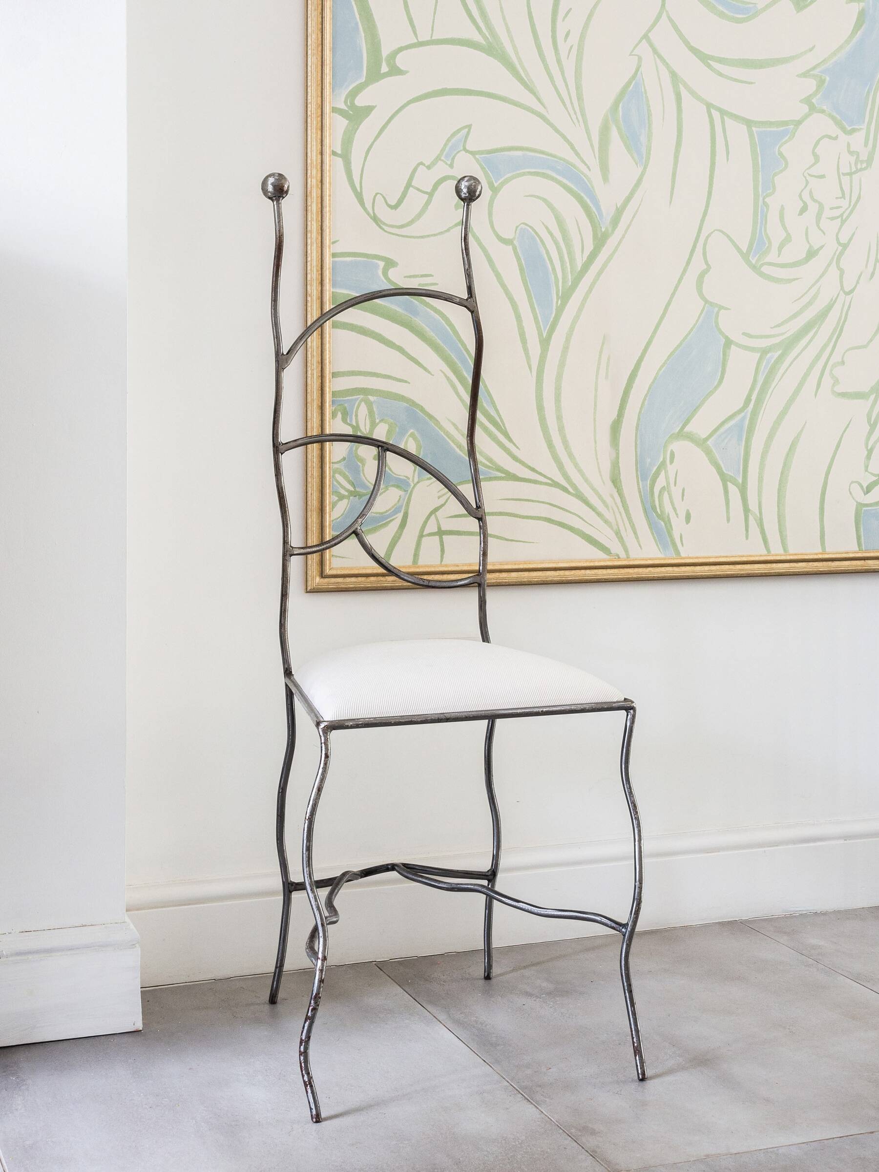 A minimalist but unique metal chair with a white cushioned seat positioned in front of a framed botanical print.