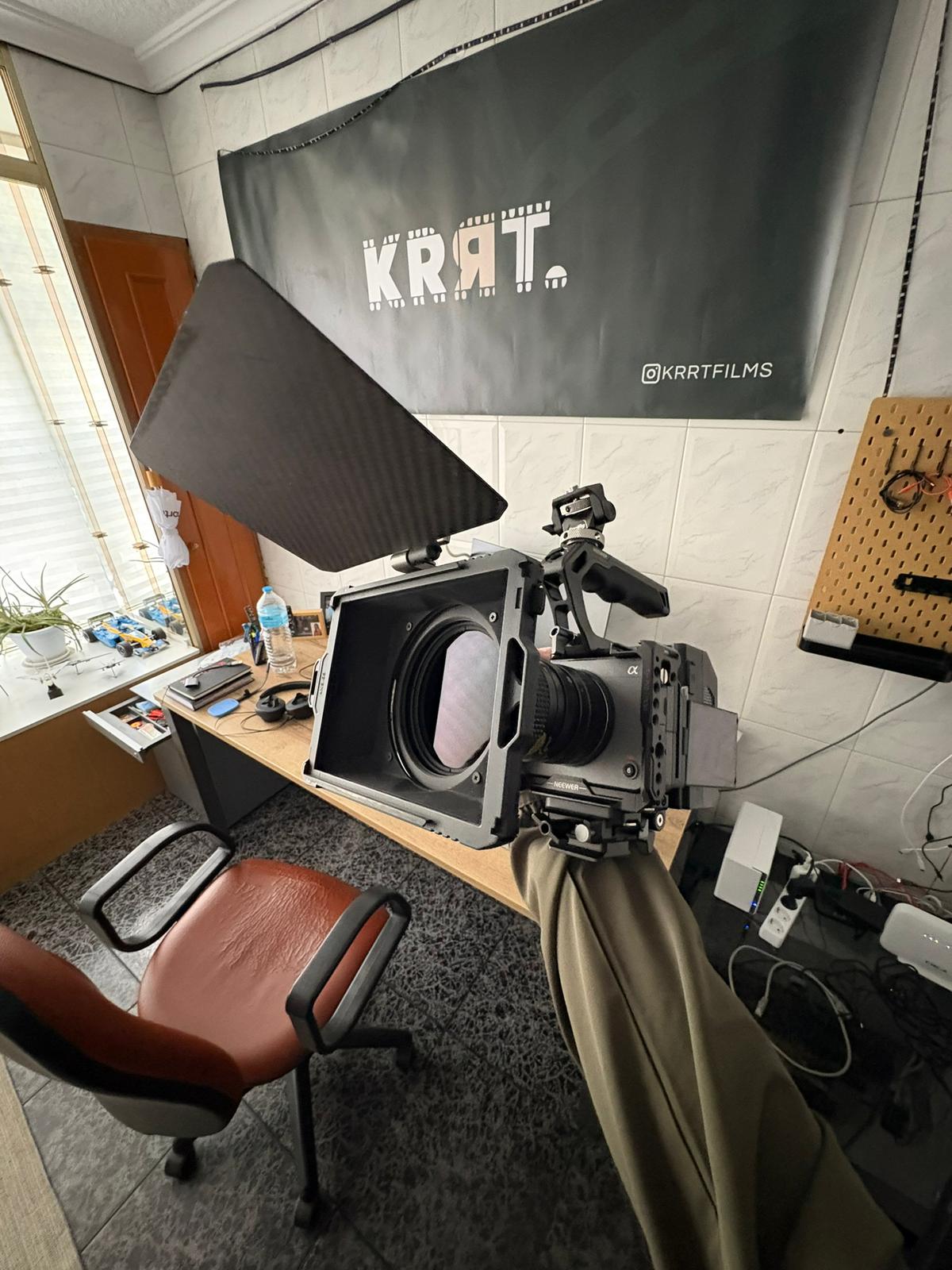 Two camera operators in a dimly lit studio adjust a large video camera. They wear headsets and caps, focusing intently. Mood is professional and focused.