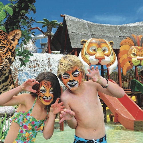 Dos niños con pintura facial de tigre posan con garras cerca de una zona de juegos acuáticos con decoraciones y toboganes temáticos de animales.