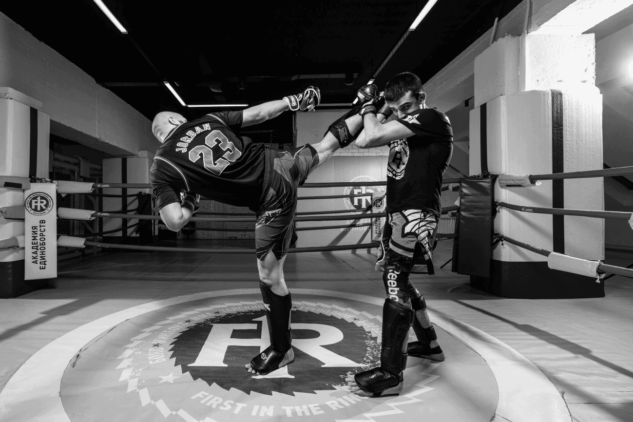 Two kickboxers sparring inside a training ring, one delivering a high roundhouse kick while the other blocks with raised gloves