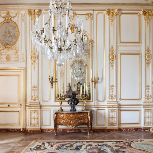Salle ornée avec des détails dorés, un grand lustre en cristal, un mur miroir et une table en bois avec un buste et des chandeliers.