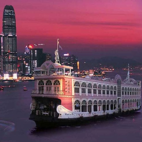 Egy nagy, díszes hajó több ablakkal úszik a vízen alkonyatkor, a háttérben egy város látképével és magas épületekkel.