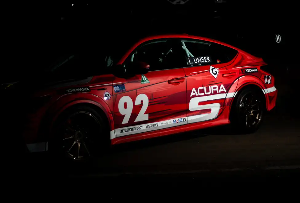 A red Acura race car emerging from the shadows at night.