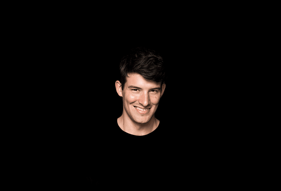 A young man with short dark hair and light skin is shown against a solid black background. He is smiling and looking directly at the camera. He wears a black crew-neck shirt.