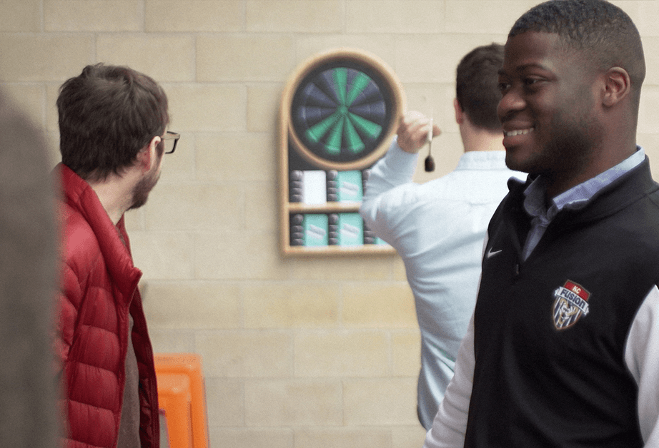 A hand holds a white card with questions for fathers about parenting experiences, positioned in front of a dartboard with teal and black sections.