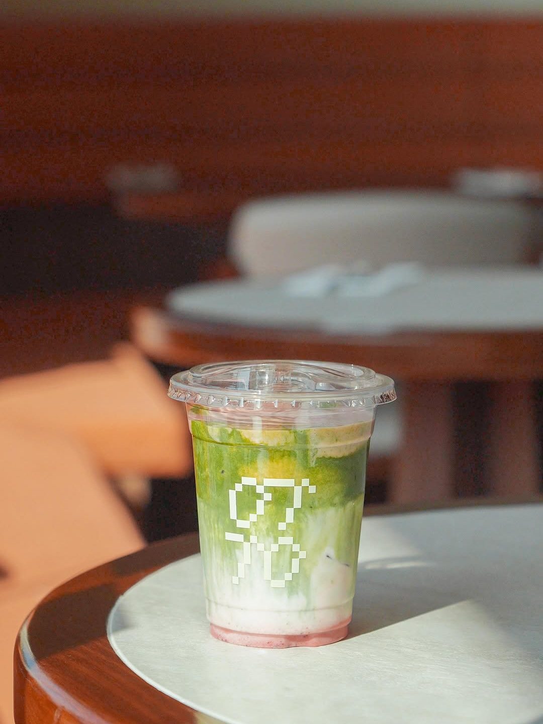 A clear plastic cup filled with a layered beverage, featuring green matcha at the top, a creamy white middle layer, and a red bottom layer, placed on a wooden table in a softly lit room.