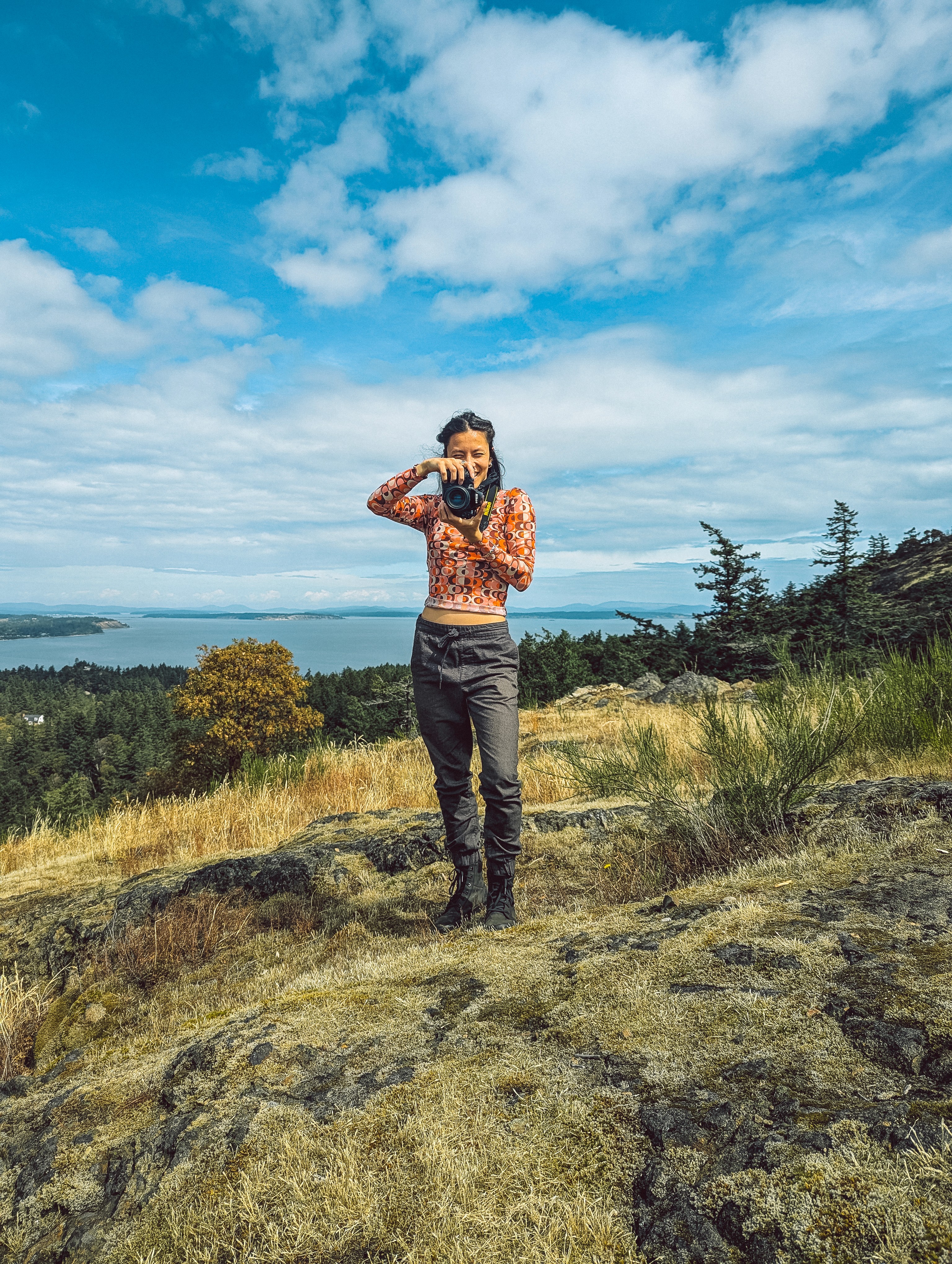 Woman taking photo on a rocky, grassy hillside overlooking water.
