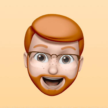 3D emoji-style avatar of a smiling man with red hair, a full beard, glasses, and blue eyes, set against a soft peach gradient background. The character has a cheerful, animated expression with an open mouth and raised eyebrows.