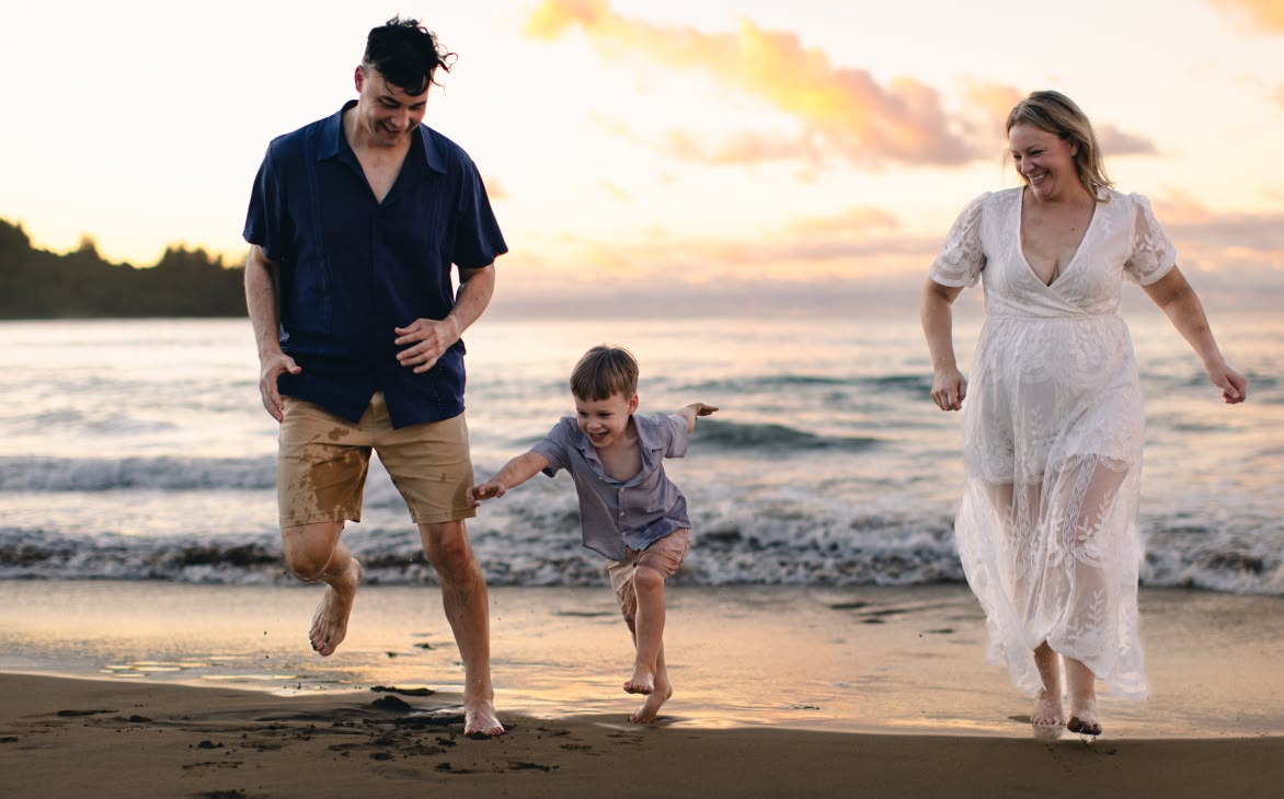 A joyful family scene on a beach at sunset, featuring an adult in a navy shirt and tan shorts, another in a white lace dress, and child in a blue shirt. They run barefoot on the wet sand near the ocean, with gentle waves and vibrant sky filled with soft clouds in the background, capturing a sense of happiness and togetherness.