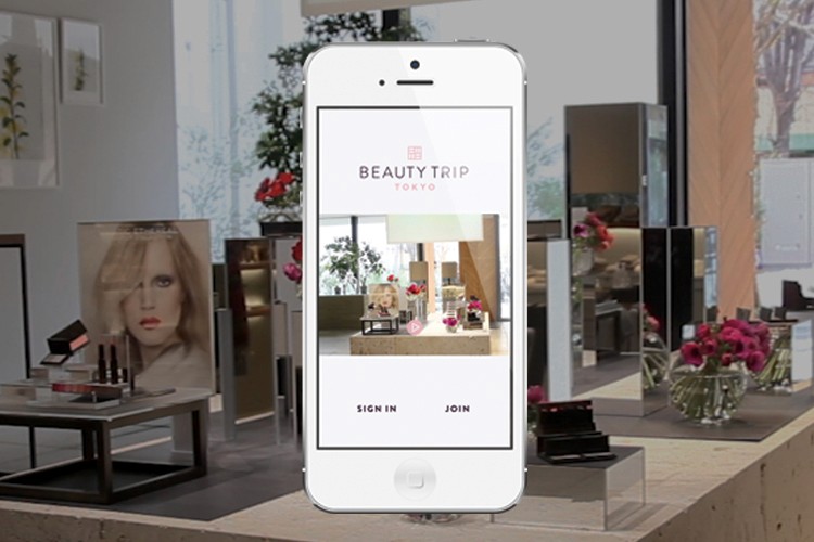 A white iPhone displays the app “BEAUTY TRIP TOKYO” on its screen. The screen shows an interior view of a store with shelves and displays. A large image of a woman’s face is visible on the left side of the screen. Below the screen, the app offers two options: “SIGN IN” and “JOIN”. The phone is positioned on a display stand with additional items.